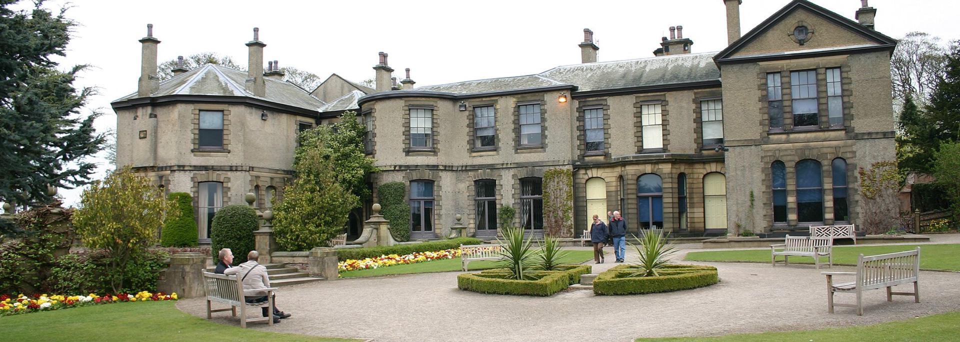 Large stone building with multiple windows, surrounded by a garden and paved area with benches.