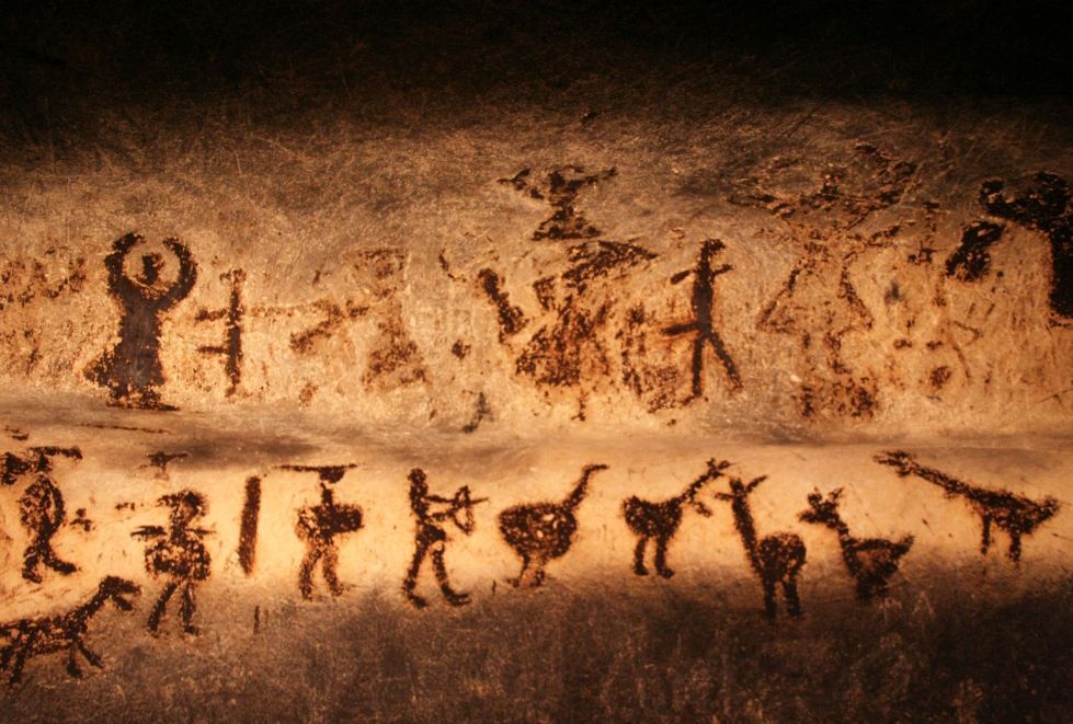 Cave painting depicting human figures, animals, and abstract symbols on a tan, rough surface.