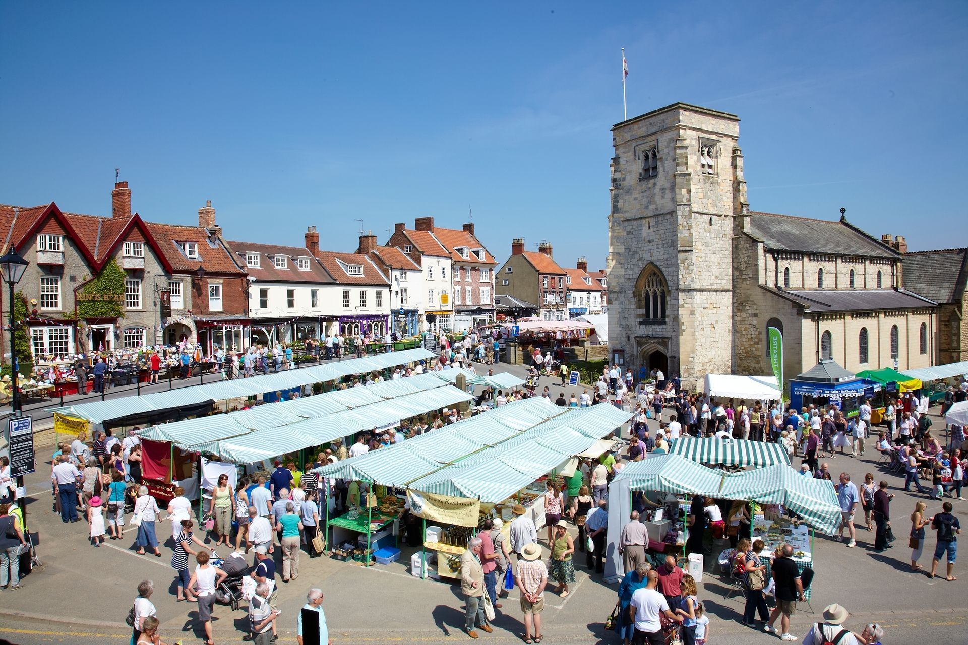 Busy market square with stalls, crowds, and a church tower on a sunny day.