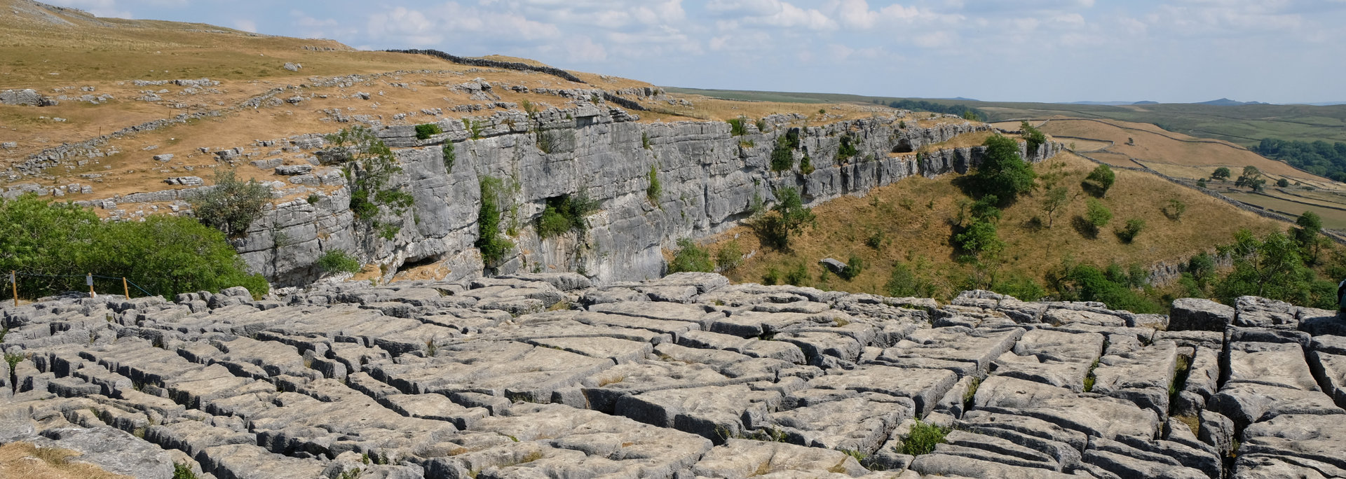 Rocky terrain with a cliff face, small trees, and a distant hillside under a blue sky.