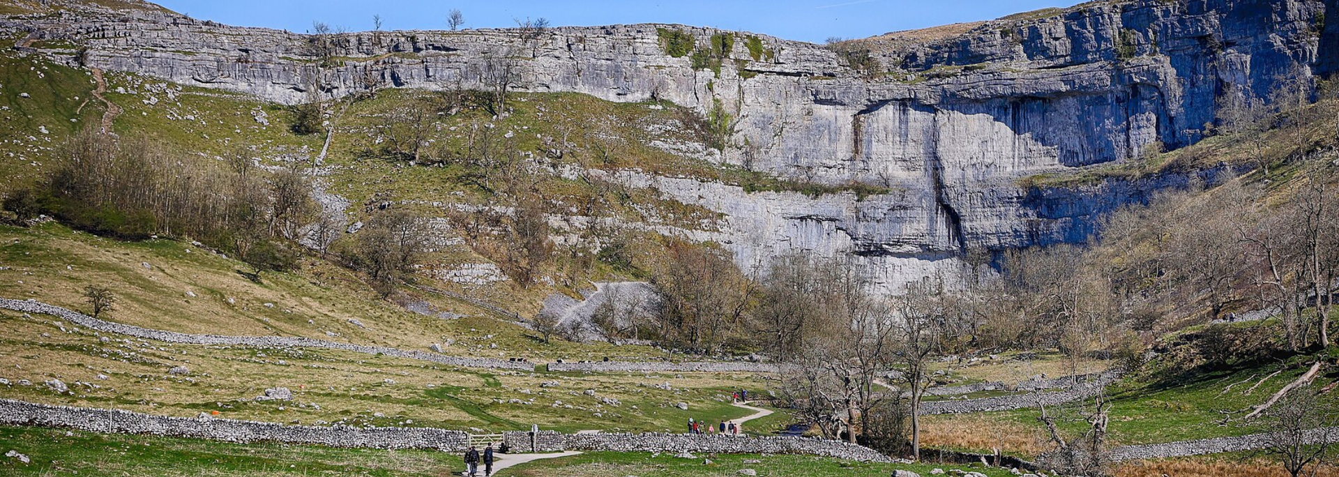 A valley with a rocky cliff face and green grassy areas, a trail and a stone wall in the foreground.