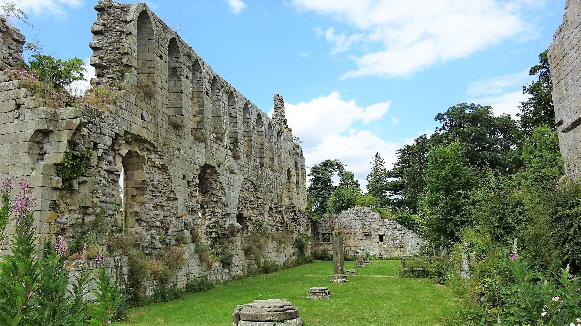Ruins of a stone building with arched windows, surrounded by green grass and trees, under a blue sky.
