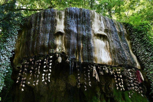 Stone structure with dripping water, resembling a face with carved eye sockets, adorned with hanging objects, surrounded by greenery.