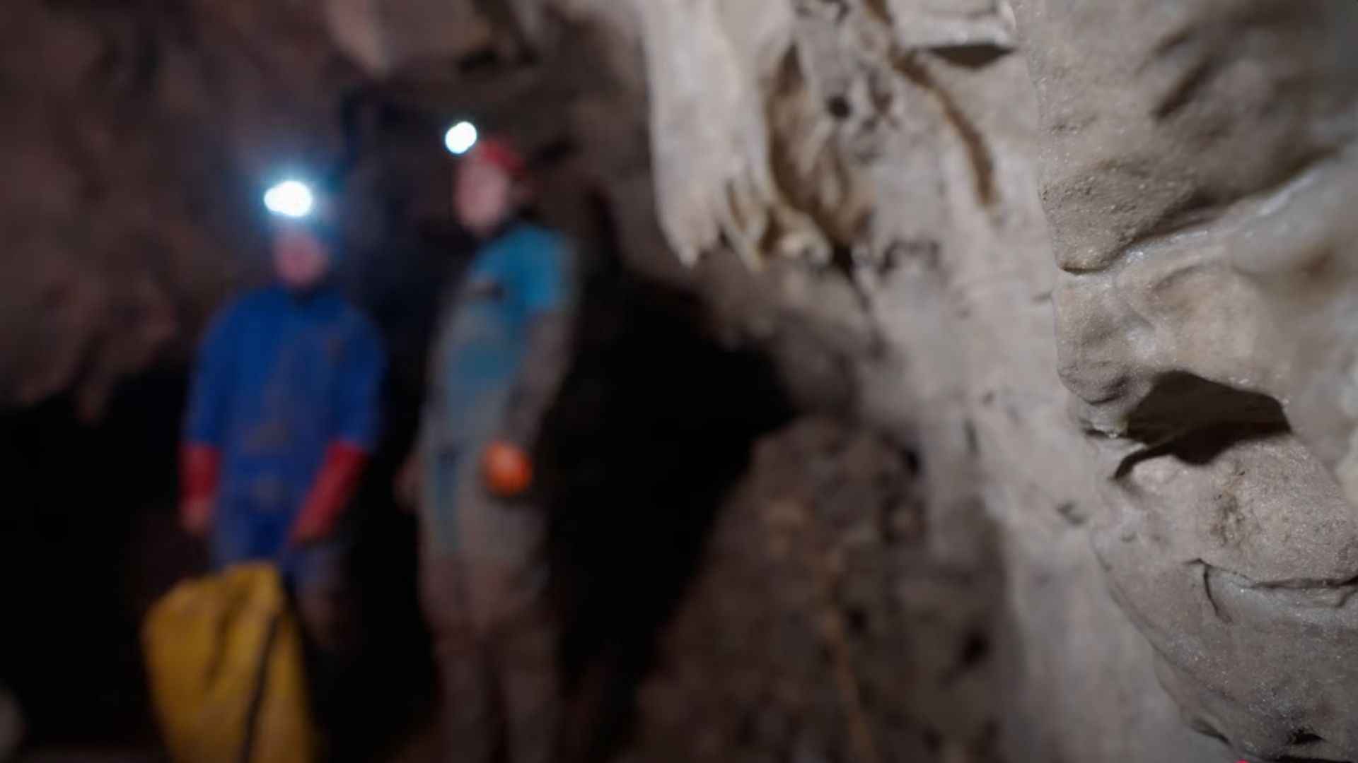 Two figures with headlamps in a cave. One in blue, the other in a light color. Cave walls are light brown and textured.
