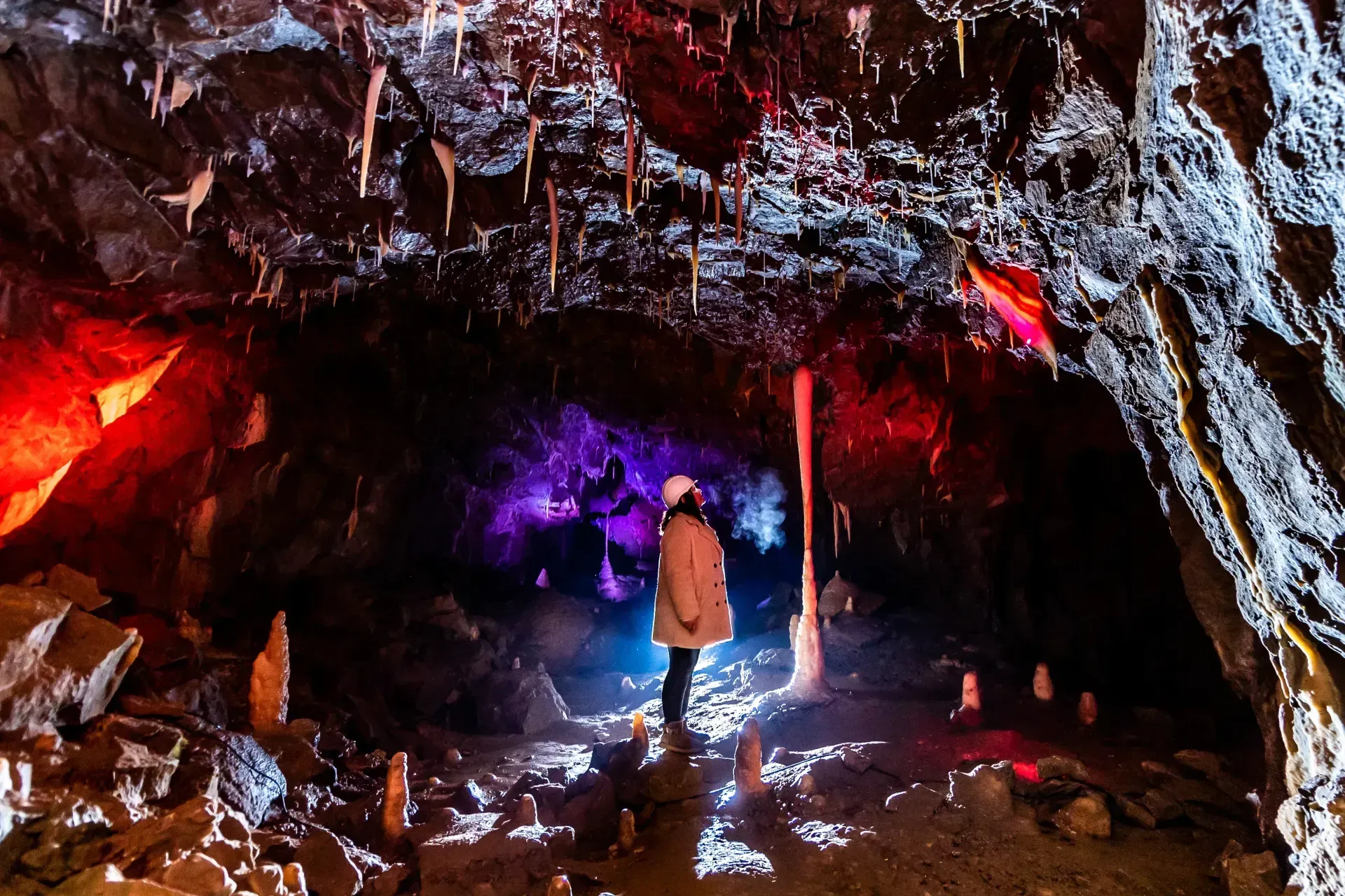 Person standing in a colorful cave illuminated by red, purple, and white lights, with stalactites hanging from the ceiling.