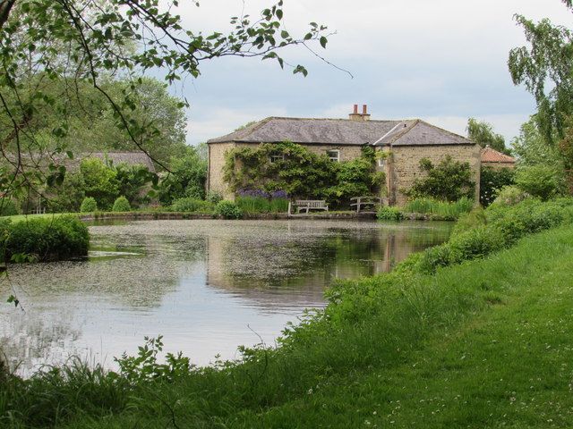 A stone building with a pond, surrounded by greenery under a cloudy sky.