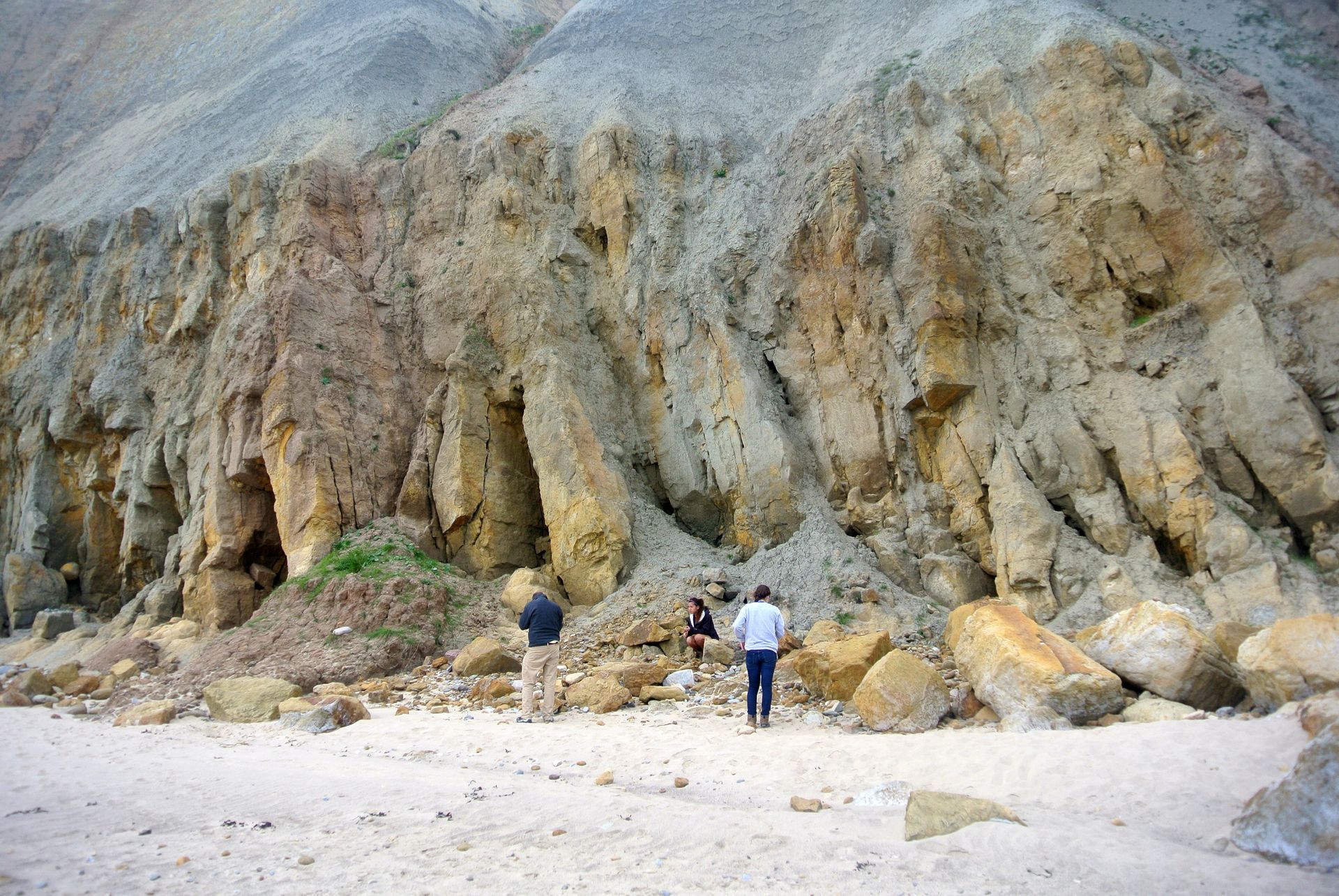 People stand at base of eroded cliff face, tan rock formations, pale sand and sky.