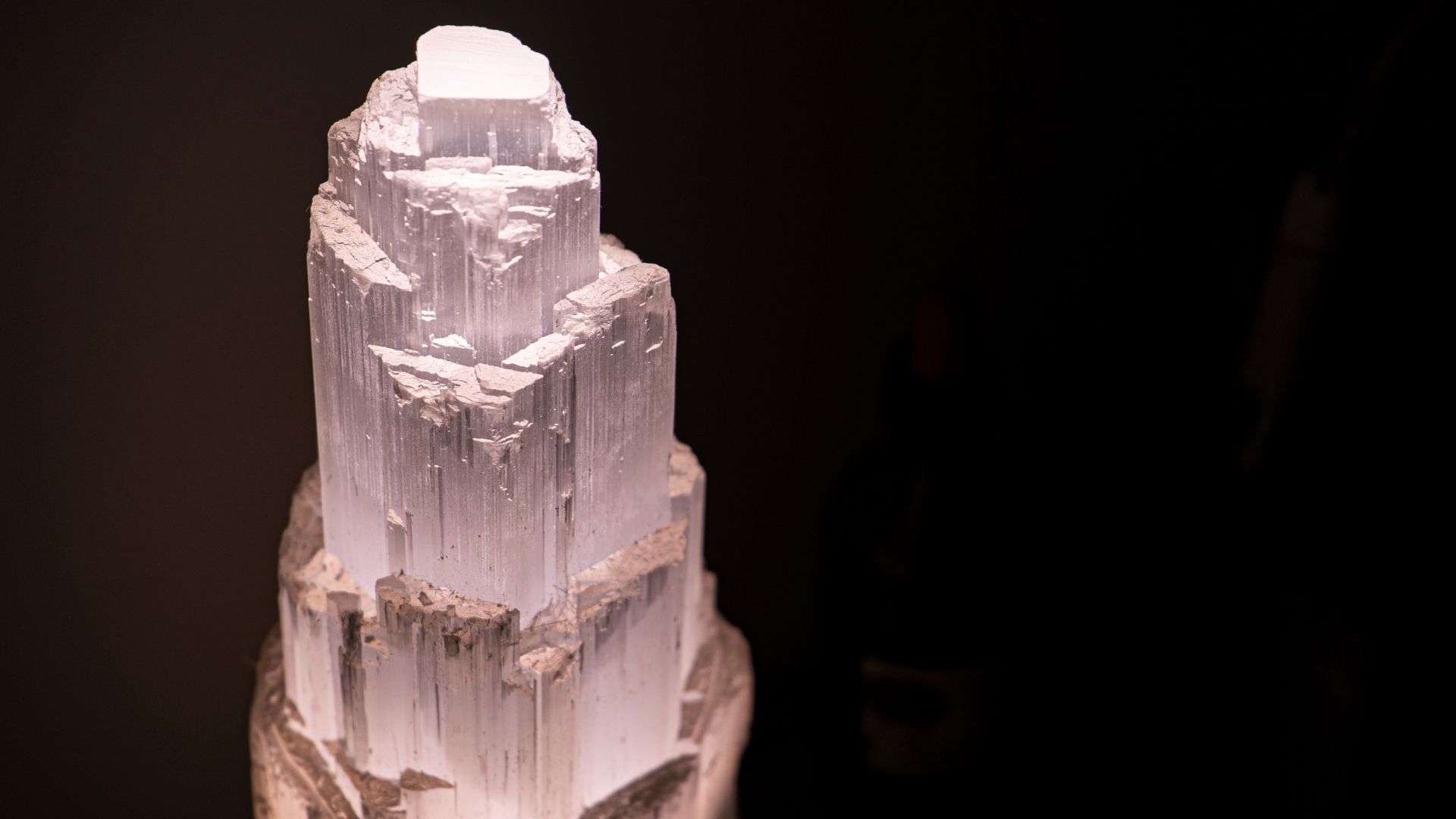 Towering selenite crystal illuminated against a dark background, showing vertical lines and layered structure.