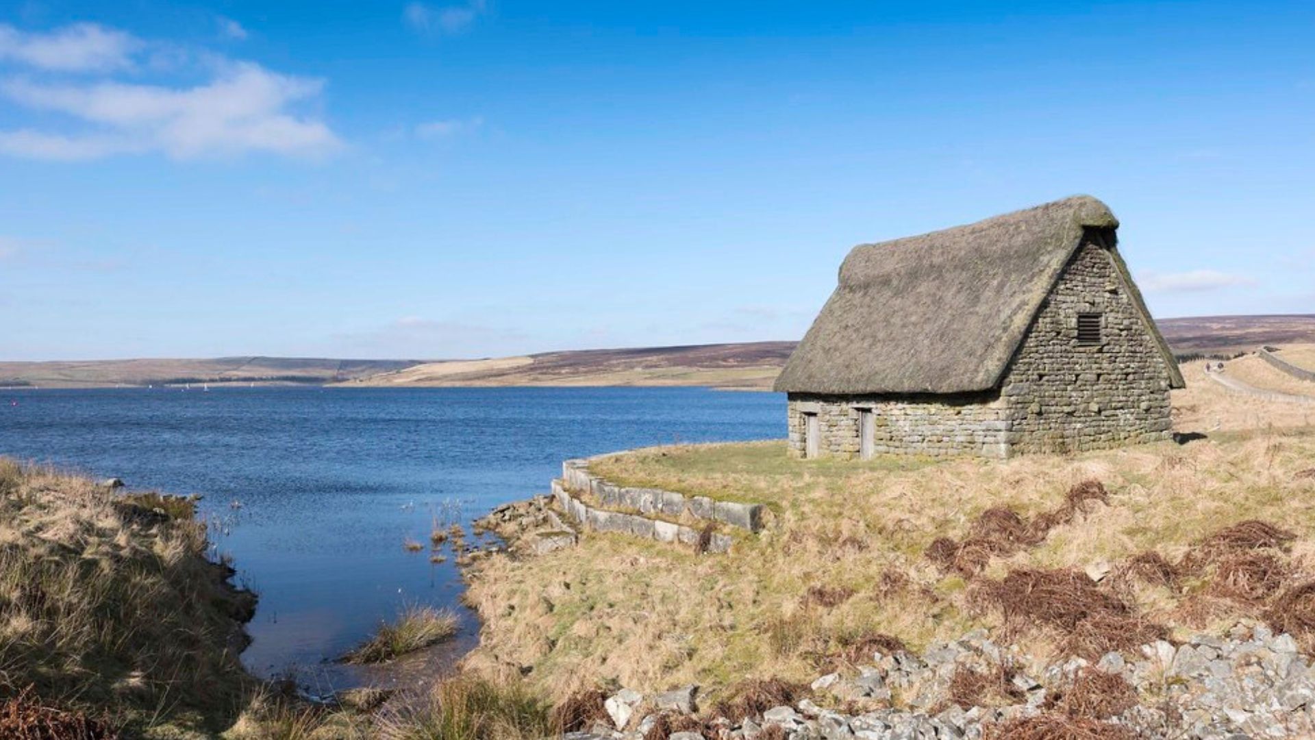 Stone cottage with thatched roof on a grassy bank beside a lake under a blue sky.