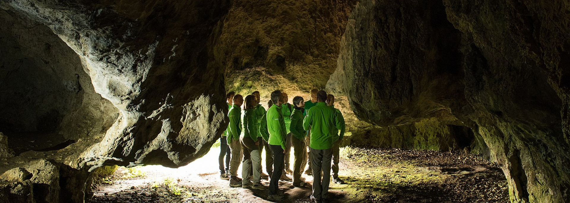 People in green jackets stand inside a dark cave, lit by a spotlight.