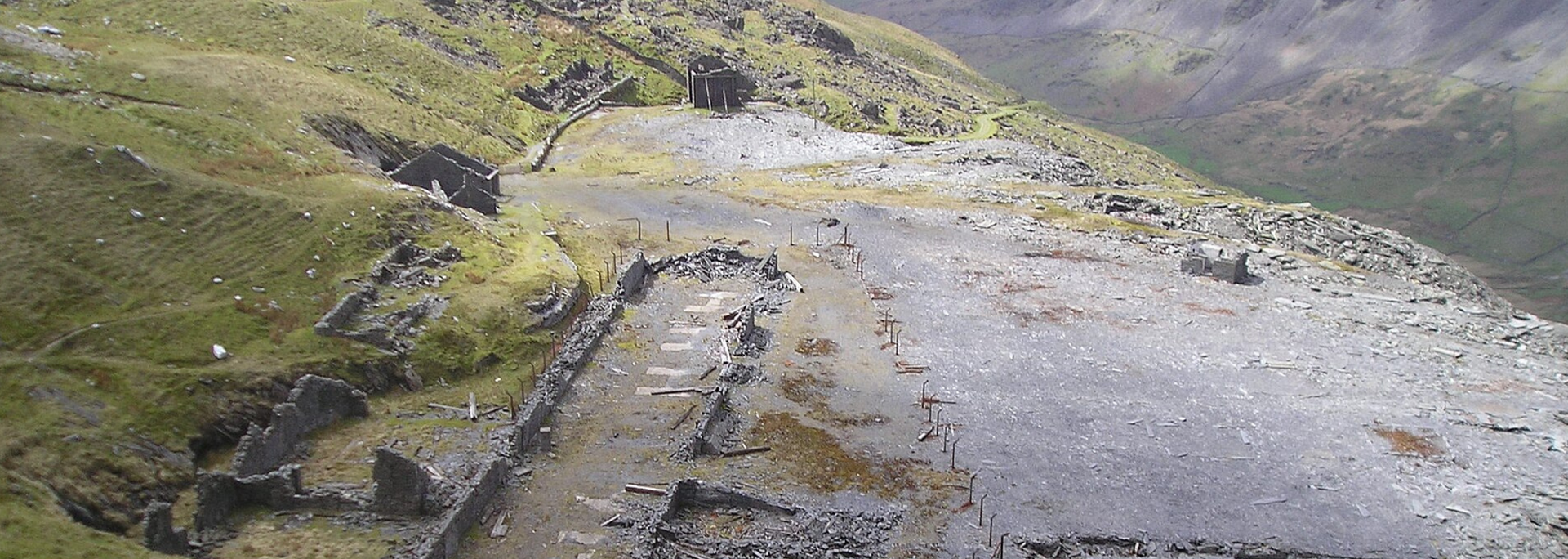 Aerial view of a mountain with a gray, rocky area and grassy slopes. Ruins are visible.