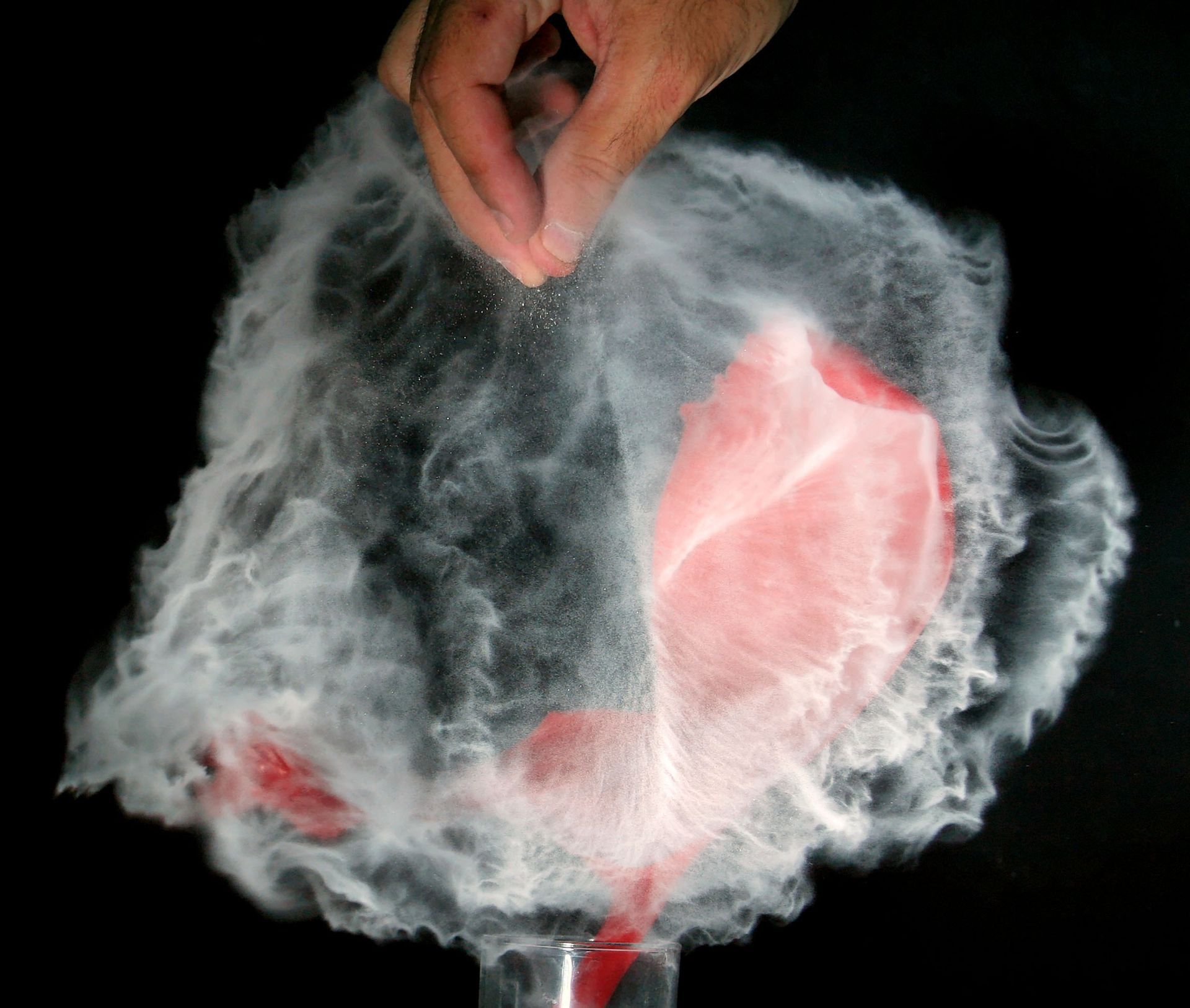 Hand sprinkles powder over a red balloon surrounded by a cloud of white powder, against a black background.