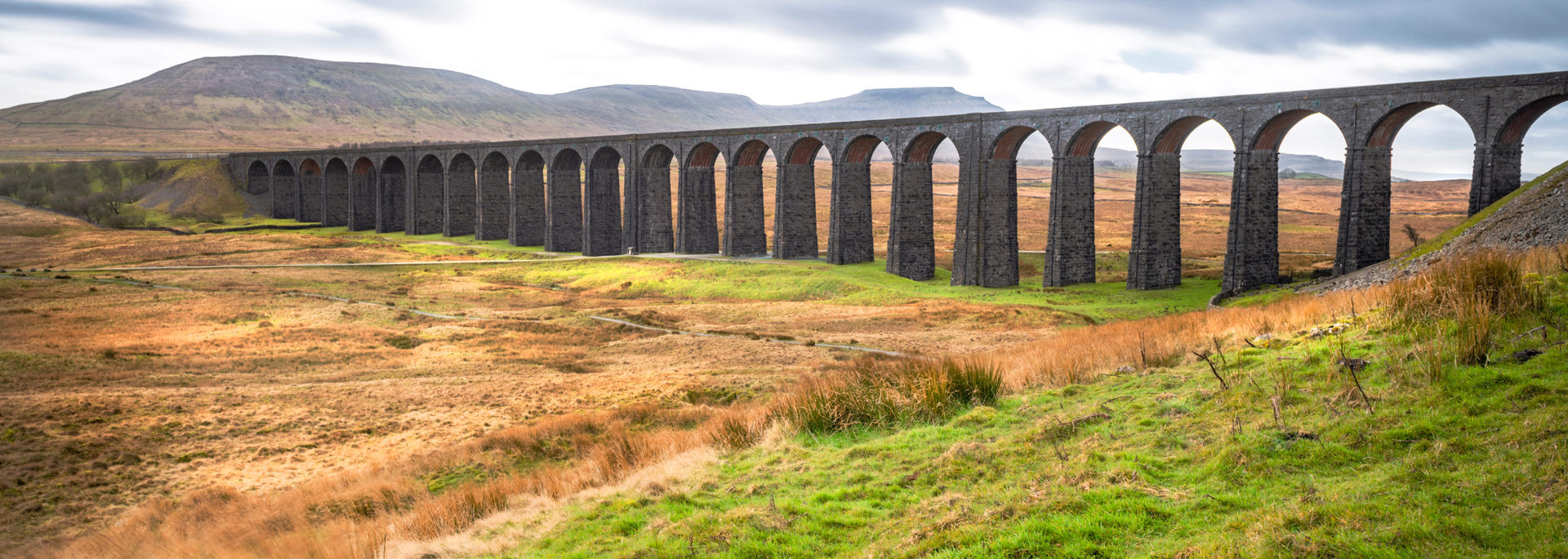 Stone arch viaduct stretches across a grassy valley under a cloudy sky.