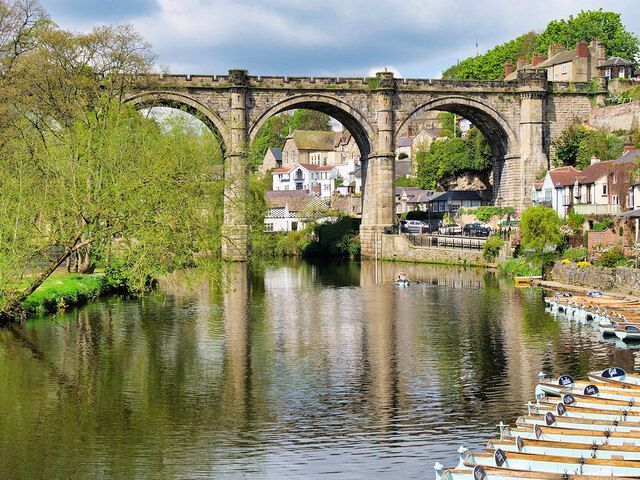 Stone arch bridge over a calm river, reflecting the structure. Green trees and buildings line the banks.