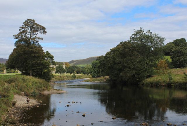 River flowing through a green landscape with trees and distant buildings under a cloudy sky.