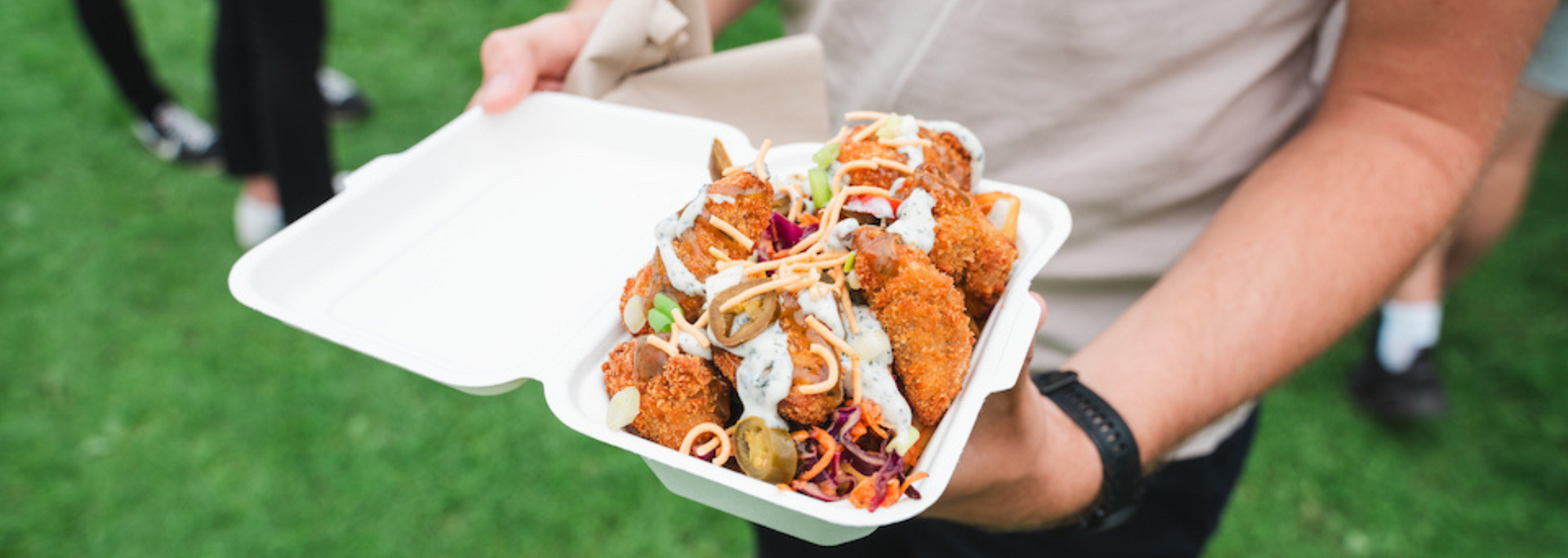 Person holding a container of fried food with sauce on a green lawn.