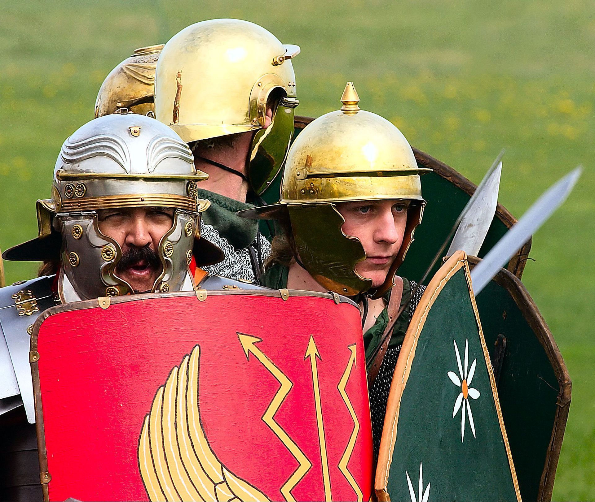Roman soldiers in helmets and armor, holding shields and swords, in an outdoor setting.