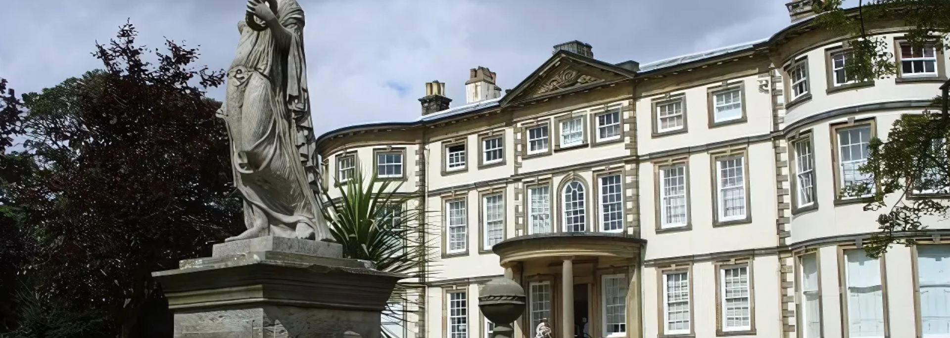 White building with multiple windows; statue in front, surrounded by trees.