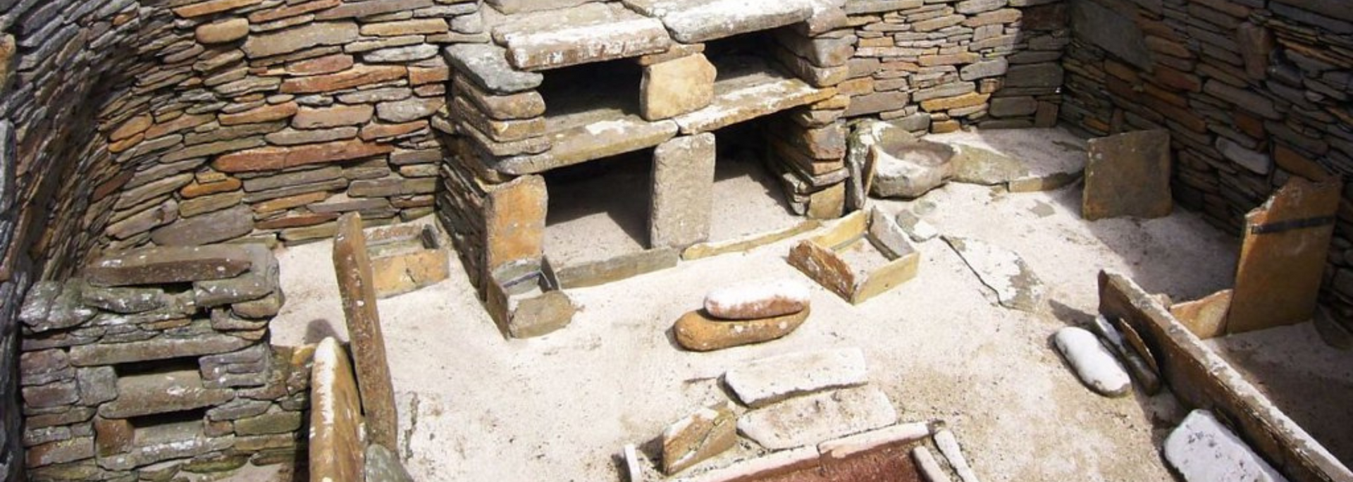 Interior view of a stone structure with shelves and compartments. Sand colored floor with scattered artifacts.