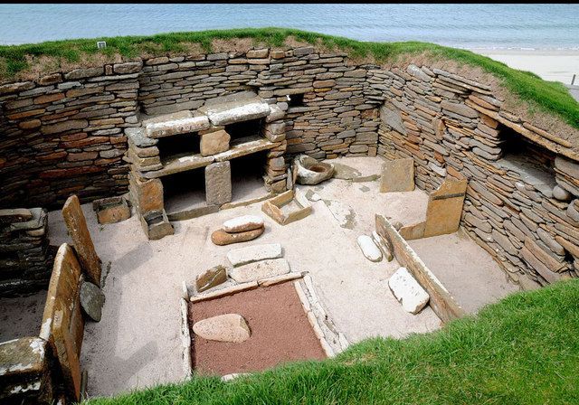 Stone-walled ancient dwelling, interior view with built-in furniture and sand floor, grass-covered exterior walls.