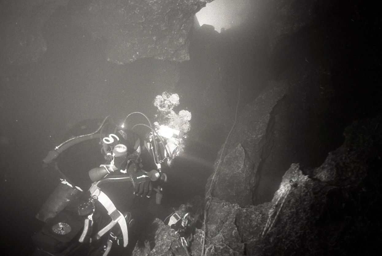 Scuba diver explores a dark underwater cave, illuminated by a bright light.