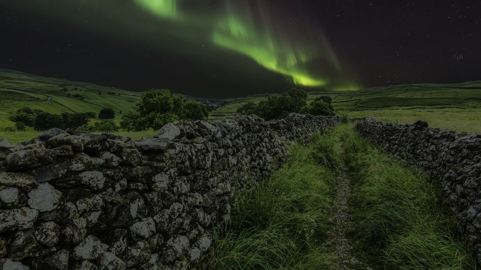 Green aurora borealis over a grassy path between dark stone walls under a starry night sky.