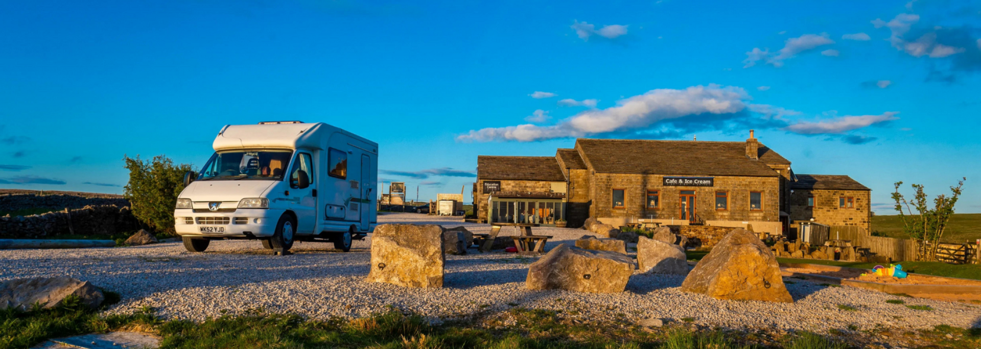 White camper van on a gravel road, approaching a stone building under a bright blue sky.