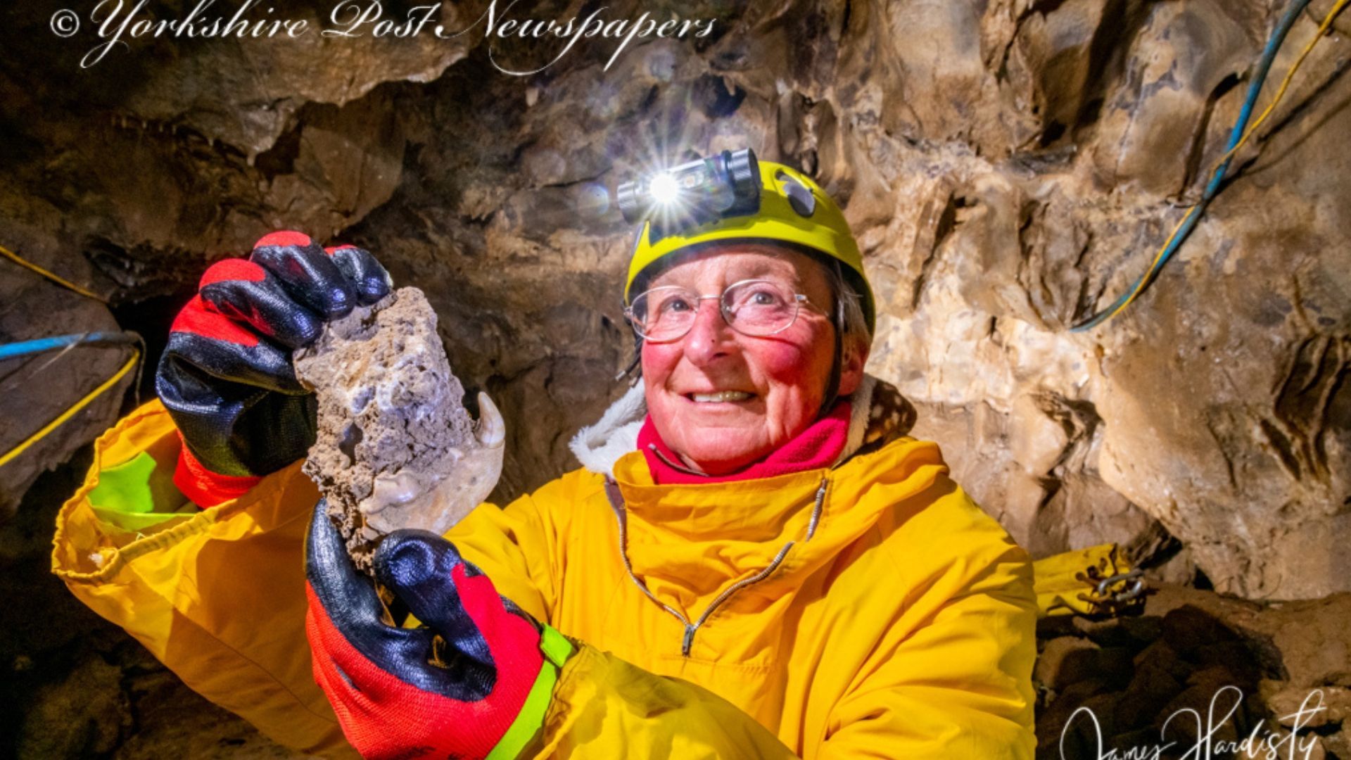Person in a cave, wearing yellow gear and headlamp, holding up a rock, smiling.