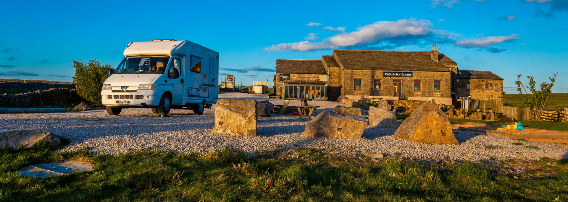 A white RV parked on a gravel road in front of a stone building with a blue sky background.
