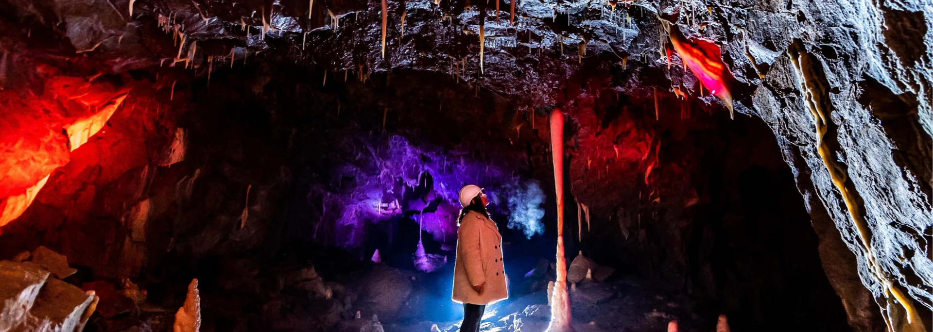 A person in a cave with colorful lights. Stalactites hang from the ceiling.