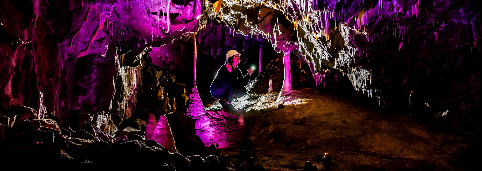 A person inside a cave with purple lighting, holding a light and looking at formations.