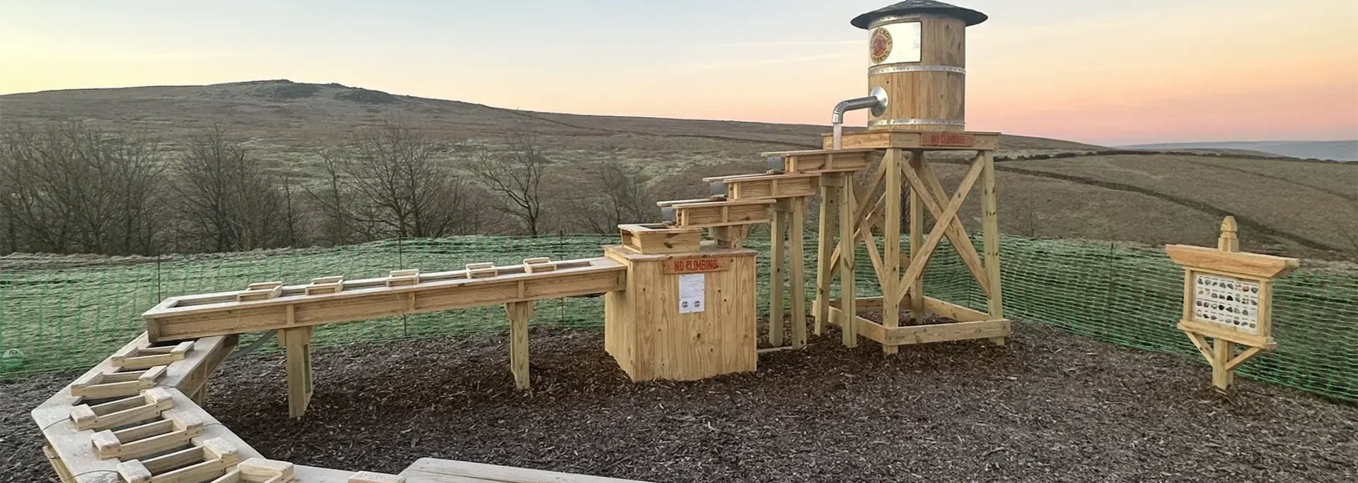 Wooden structure on a field with a tiered water feature and lookout tower. Sunset sky in the background.