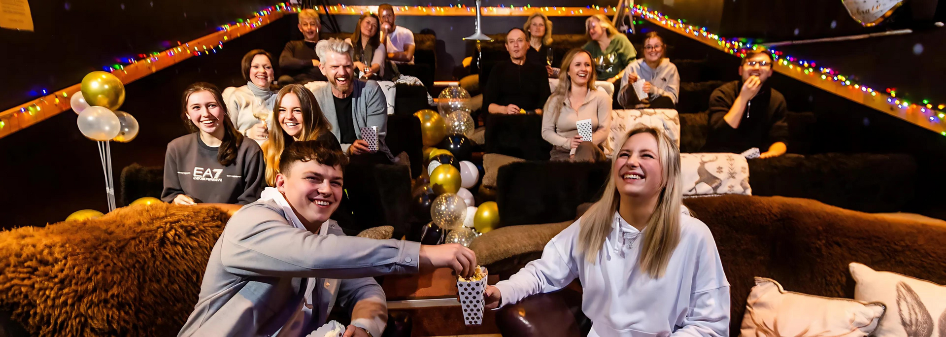 People in tiered seating, laughing. Gold balloons, fairy lights, and two people holding cups.