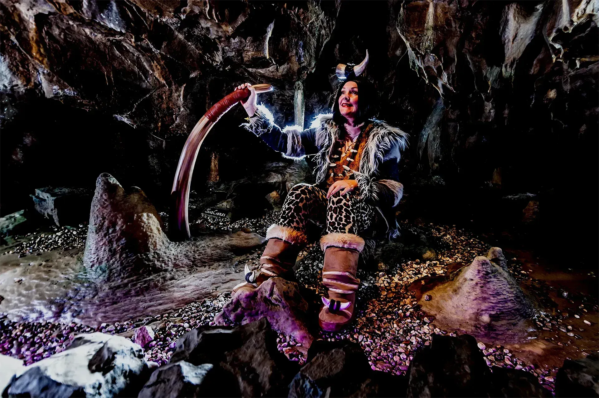 Person in horned costume holding a scythe, seated in a cave-like setting with shiny rocks.