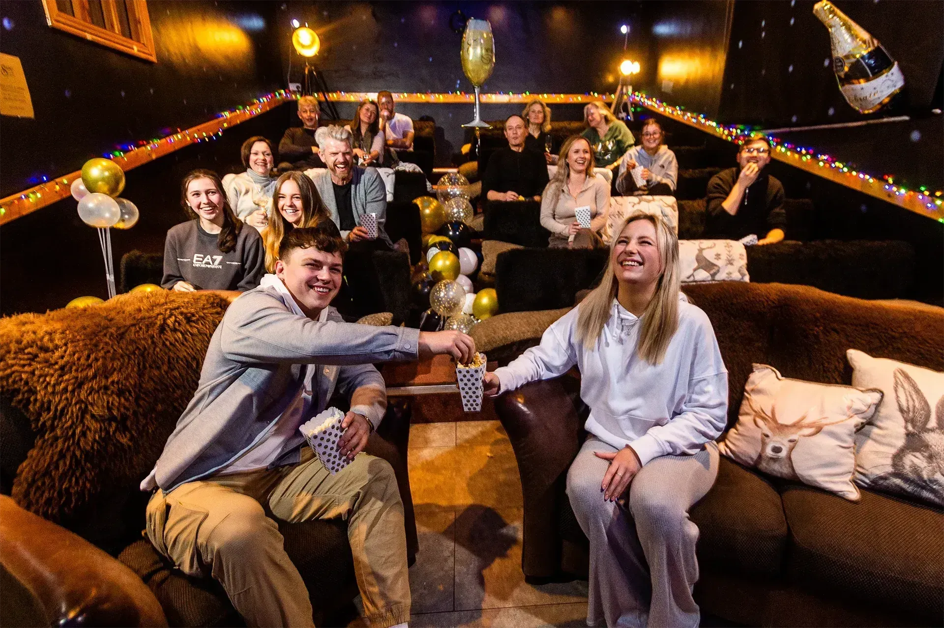 People in a home theater, raising popcorn cups. Festive balloons, string lights. Dim, cozy setting.