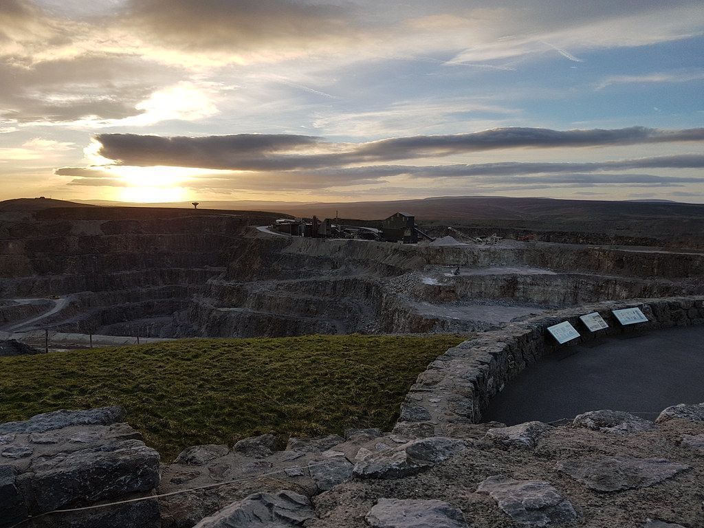 Sunset over a quarry with layered rock walls, a stone wall in the foreground, and a cloudy sky.