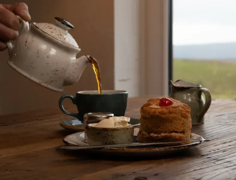 Teapot pouring tea into a cup next to a scone with clotted cream and jam.