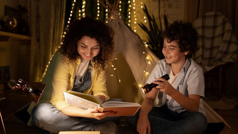 Woman and boy reading a book in a tent, lit by string lights and a flashlight.