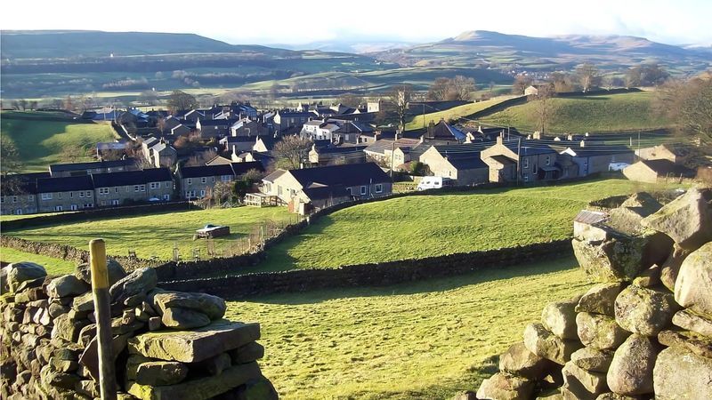 Rolling green hills overlooking a quaint village with stone buildings and fields.