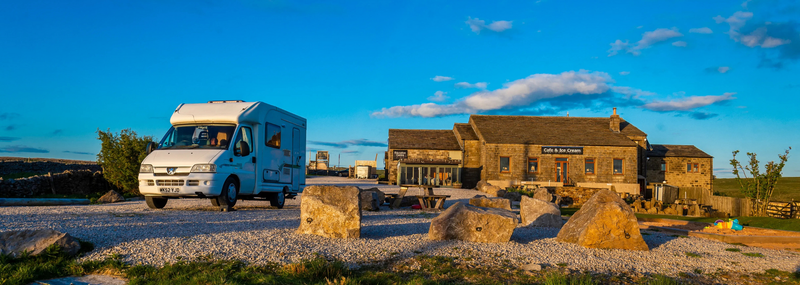 A white camper van on a gravel road, near a stone building under a bright blue sky.