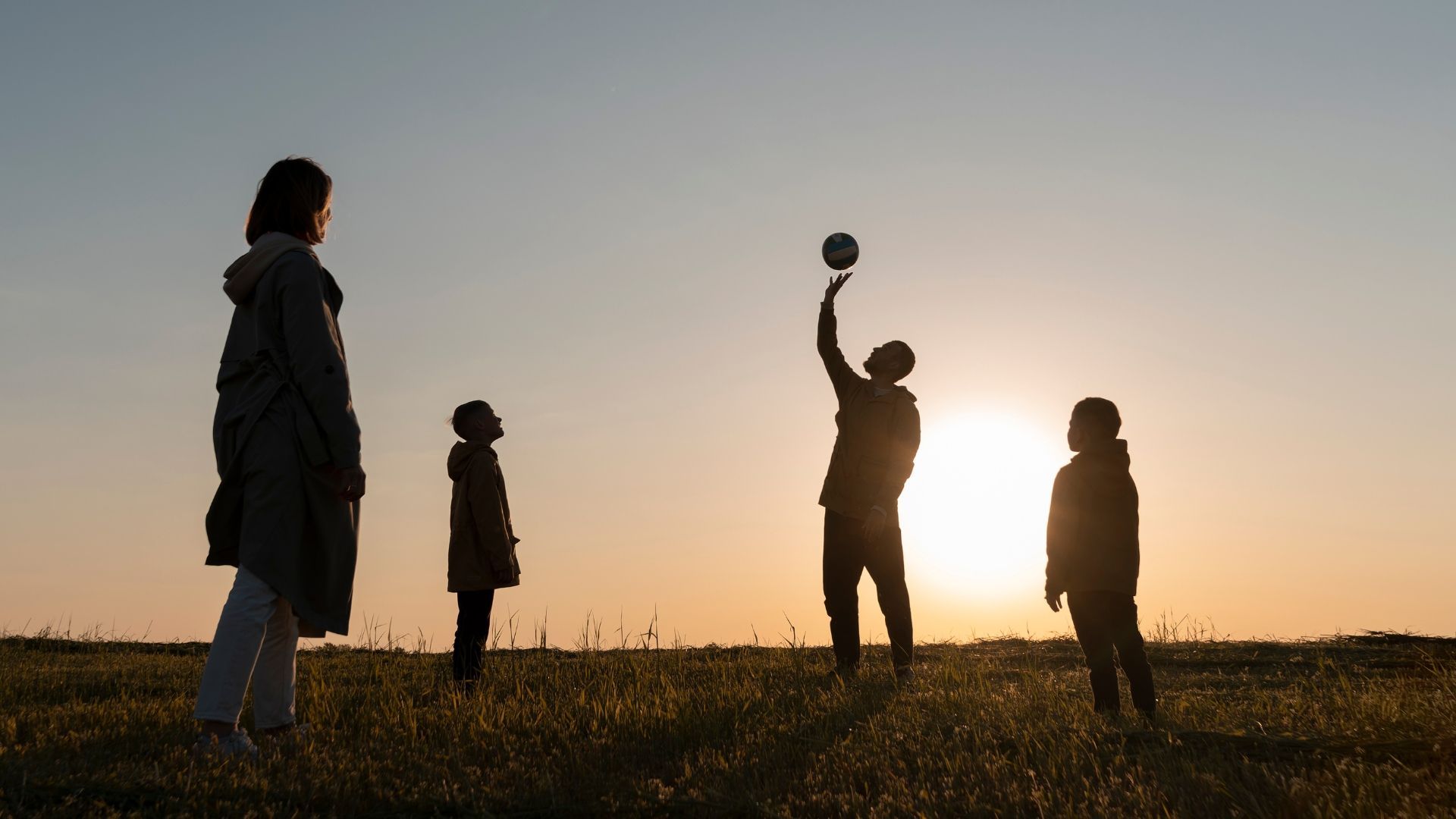 Silhouetted figures playing with a ball in a grassy field at sunset.