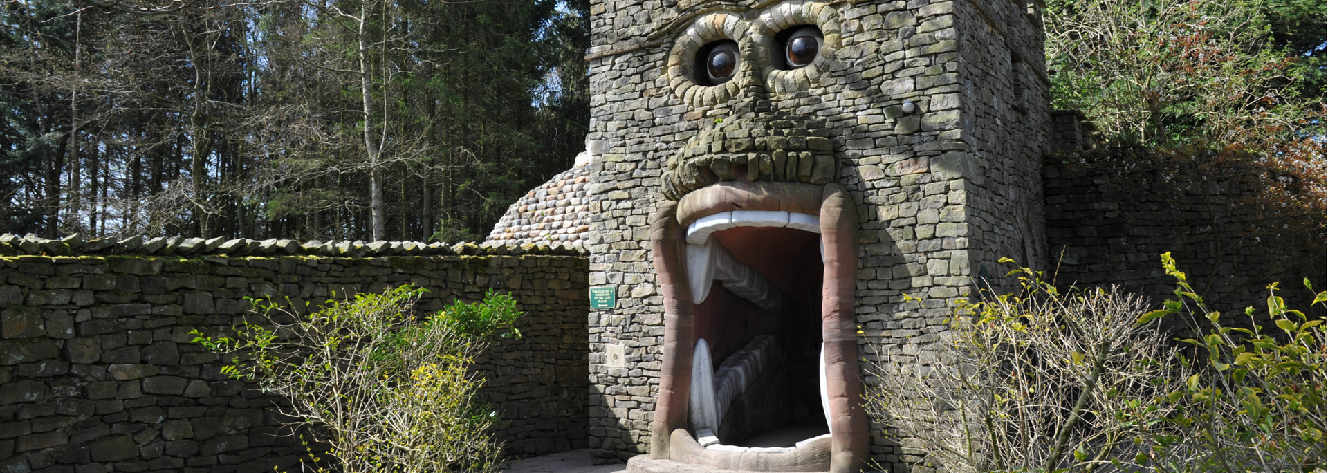 Stone structure with a large open mouth and eyes, resembling a face; surrounded by greenery and stone walls.
