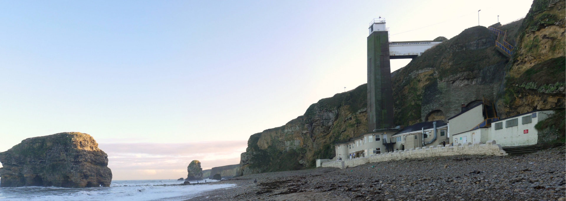 Coastal view of a rocky beach with a cliffside building and a tall tower structure; blue sky.