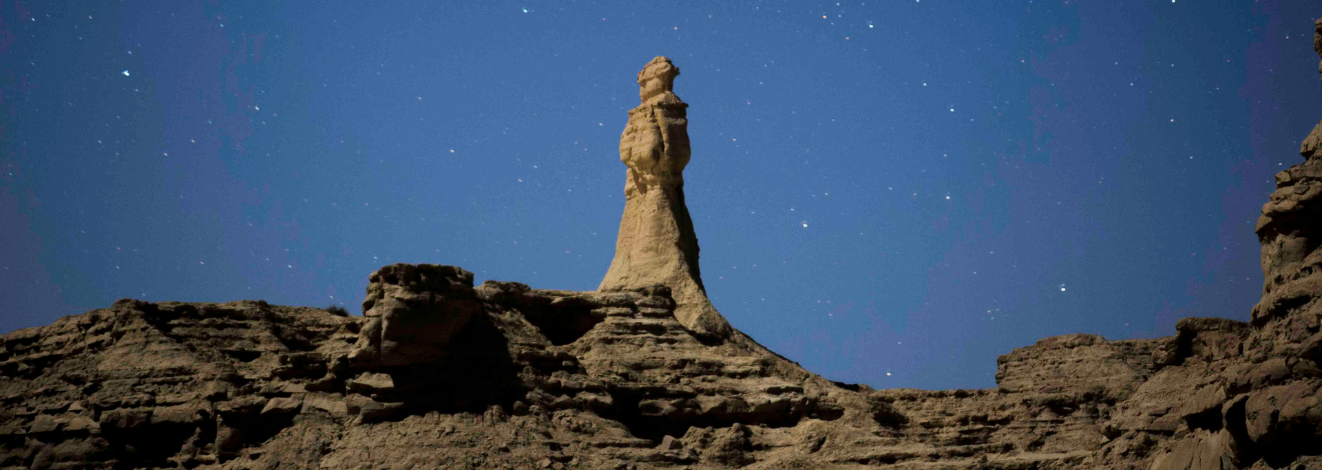 Tall sandstone rock formation under a starry night sky. The landscape is rocky and brown.