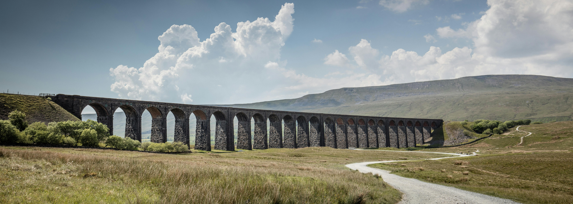 An arched stone bridge spans a grassy valley under a cloudy blue sky.