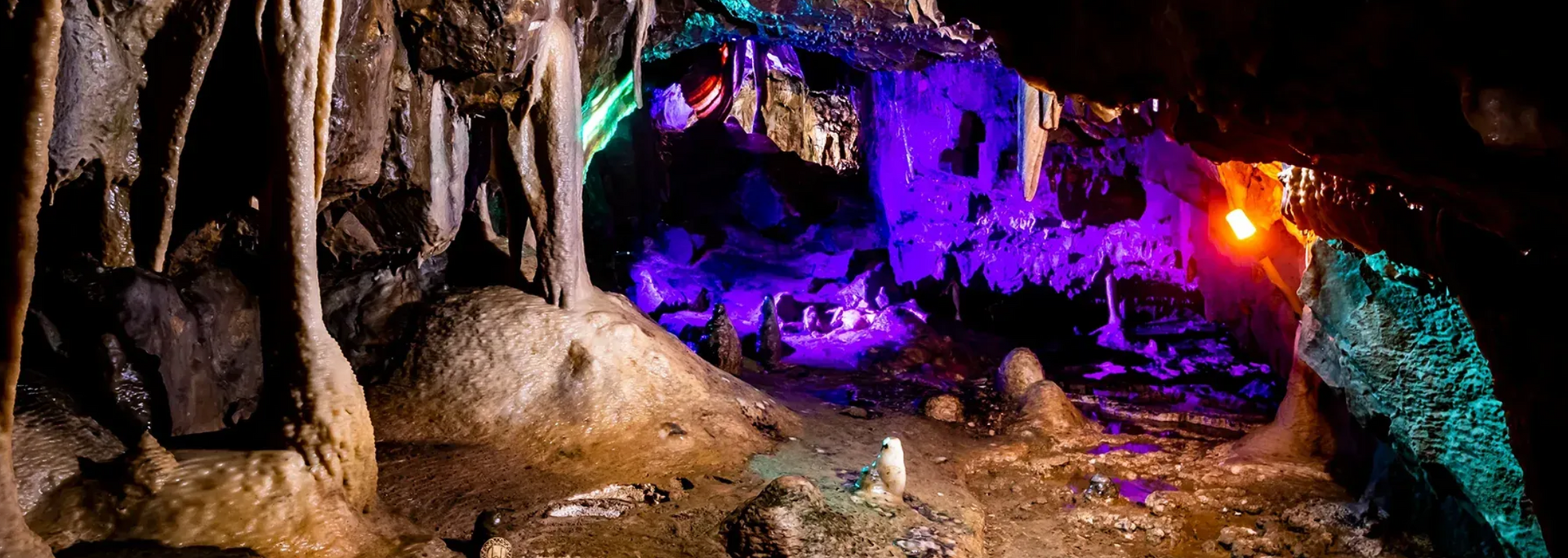 A colorful cave interior lit with purple, green, and orange lights, featuring stalactites and stalagmites.