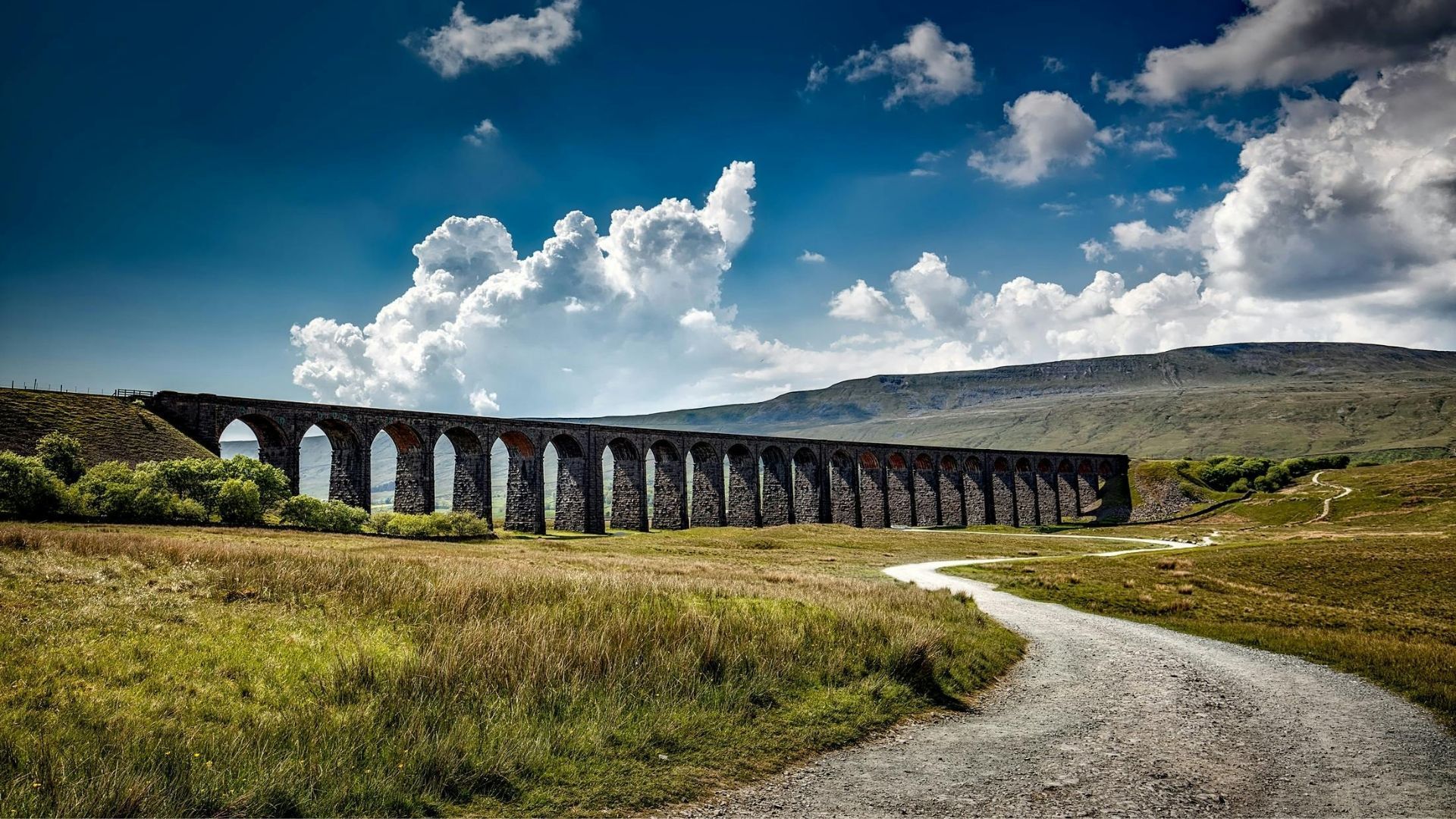 Stone viaduct arches over a grassy valley, under a cloudy blue sky.