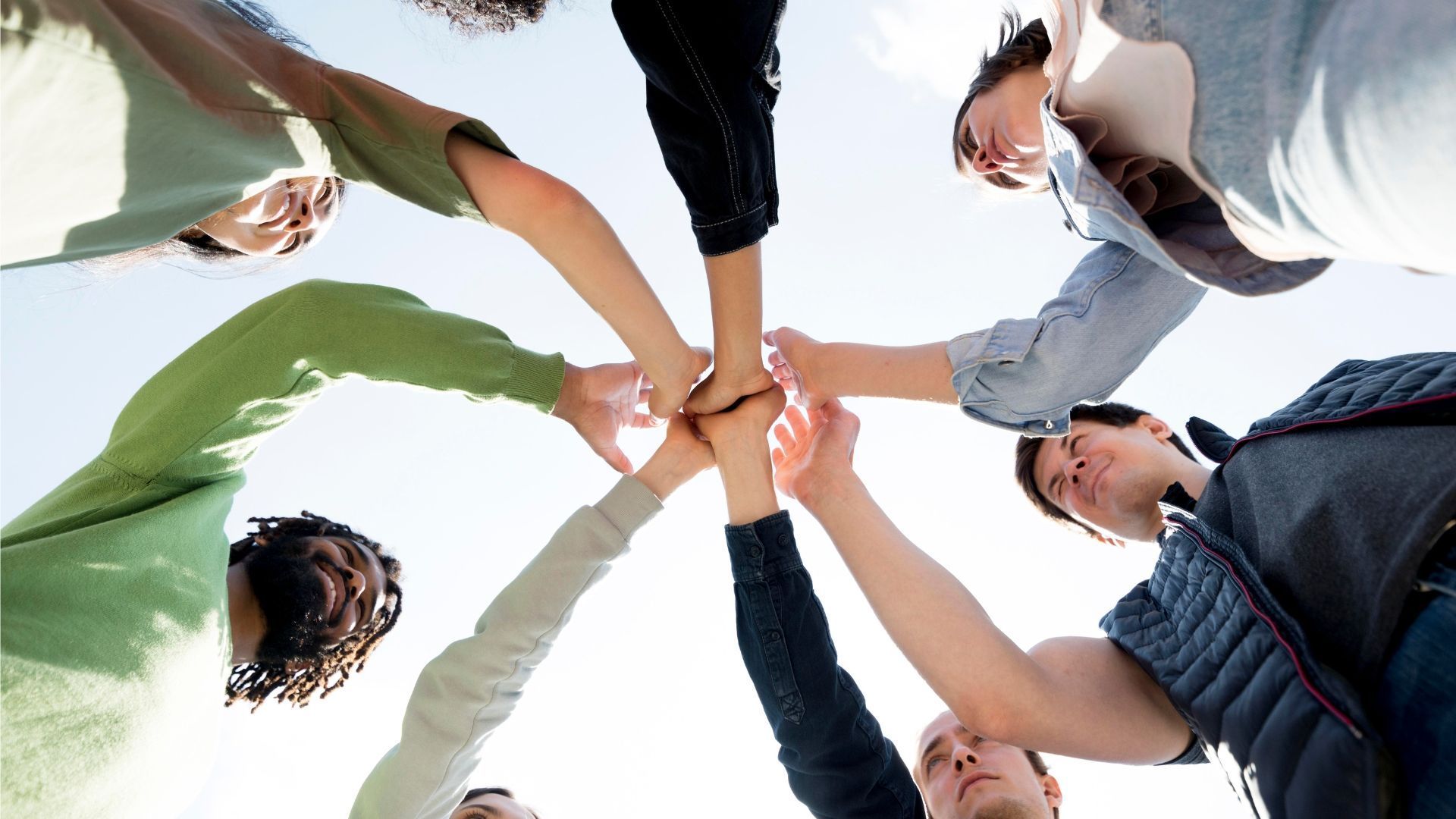 Group of people with hands joined in a circle, viewed from below against a bright sky.