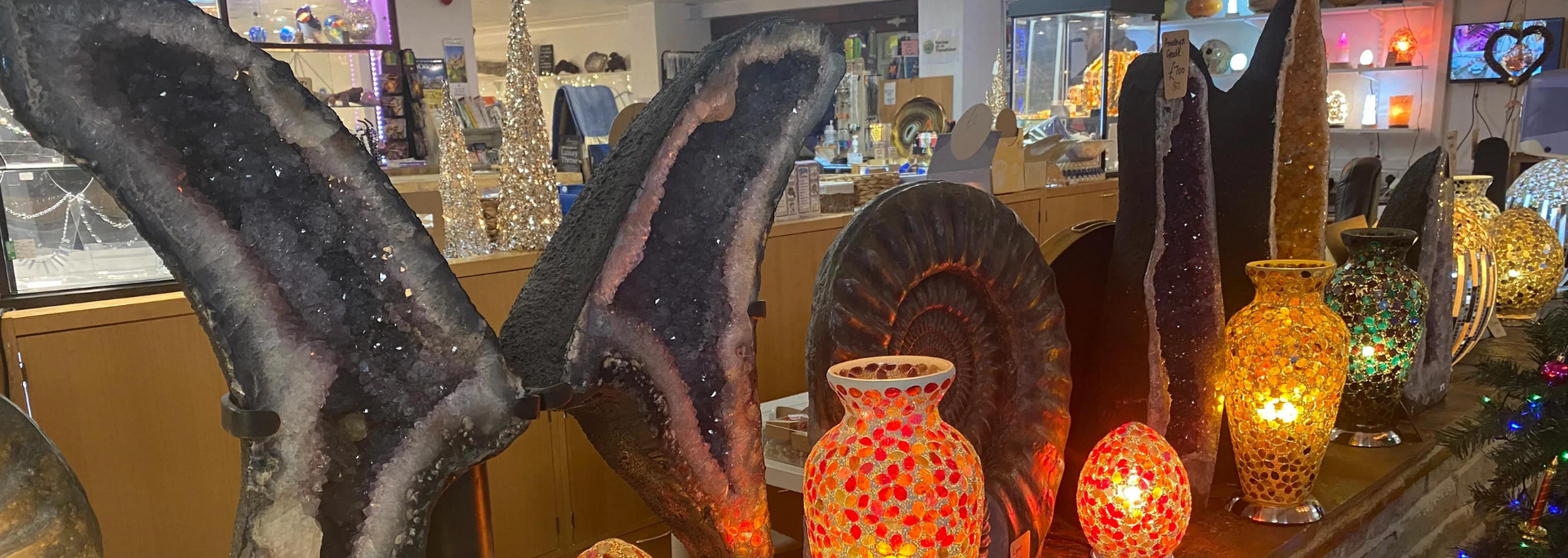 Display of crystals, lit lamps, and decorative items in a shop. Dark amethyst geodes, round brown shell.