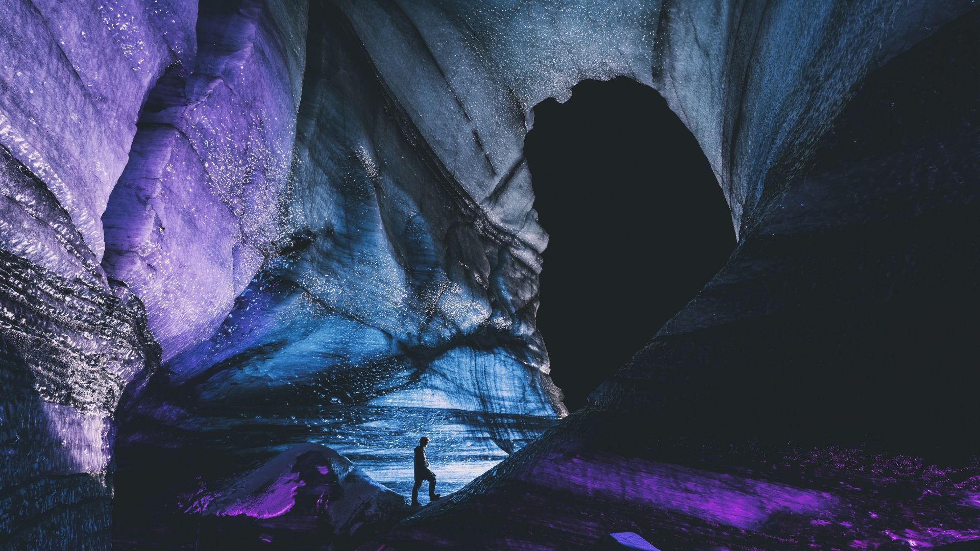 Person in dark ice cave with purple and blue glowing walls.